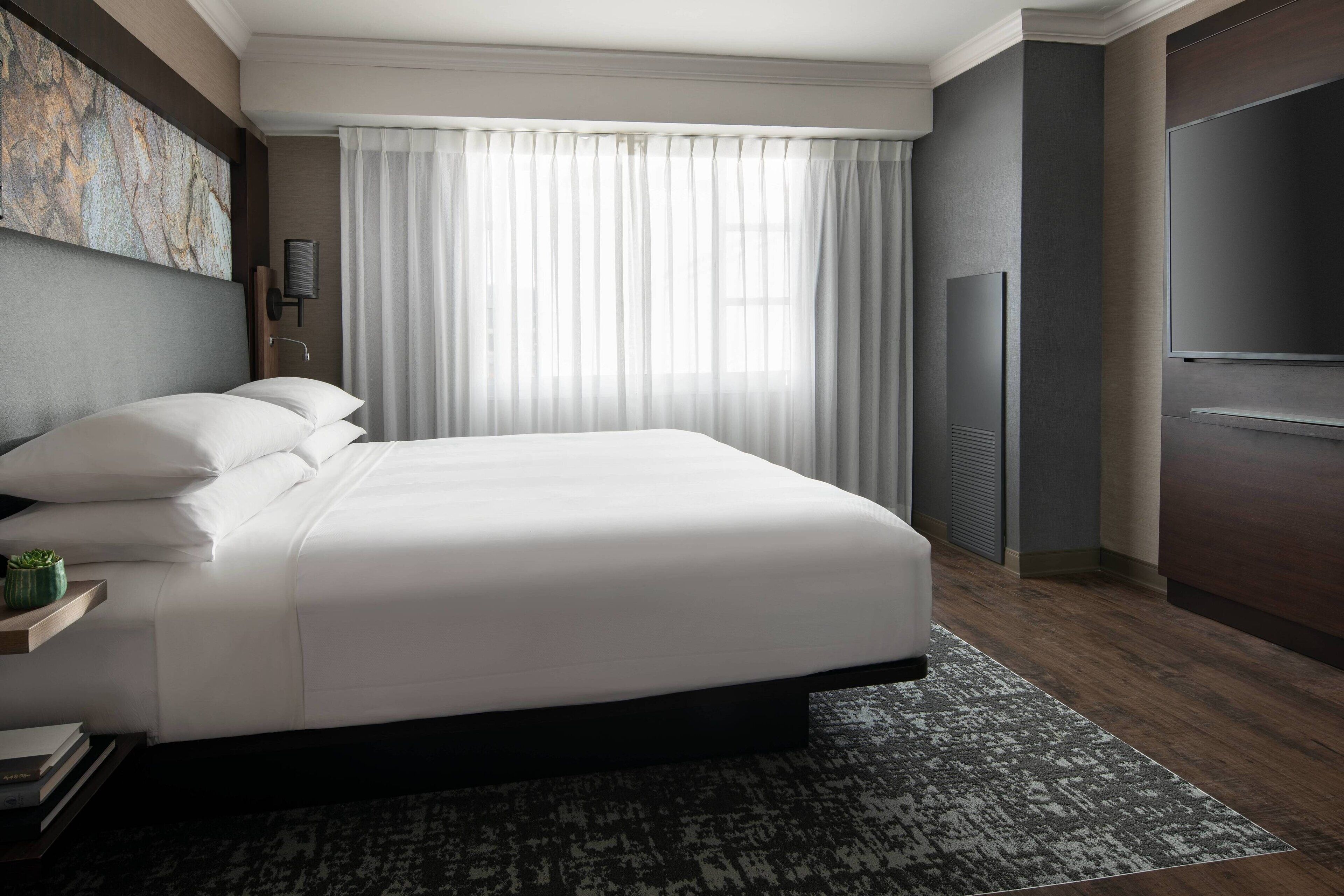 A modern hotel room with a large bed, a flat-screen TV, a large window with sheer curtains, and a dark wood floor.