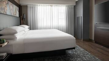 A modern hotel room with a large bed, a flat-screen TV, a large window with sheer curtains, and a dark wood floor.