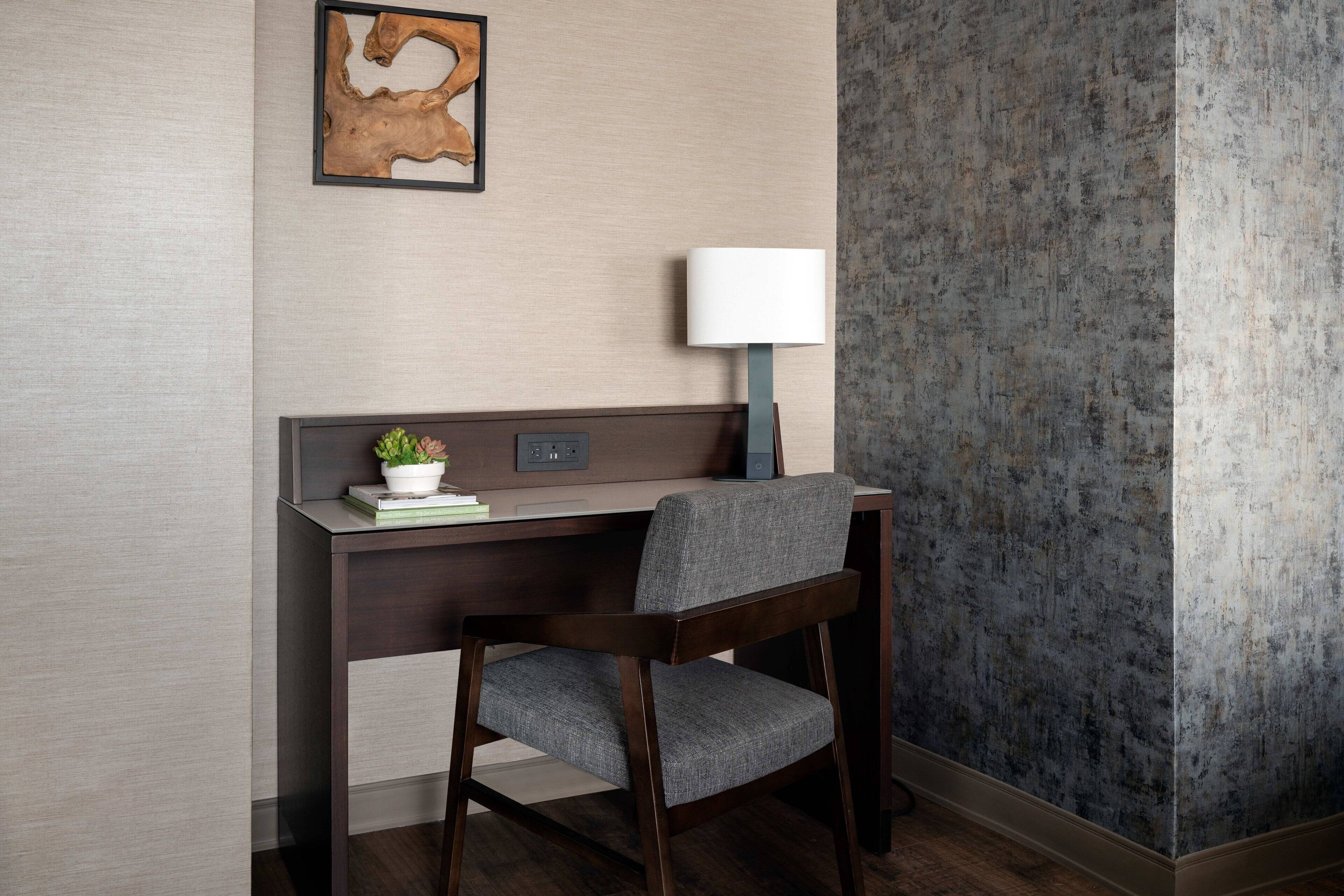 Een hotelkamer met een houten bureau, een stoel, een lamp en een kunstwerk in een lijst aan de muur.
