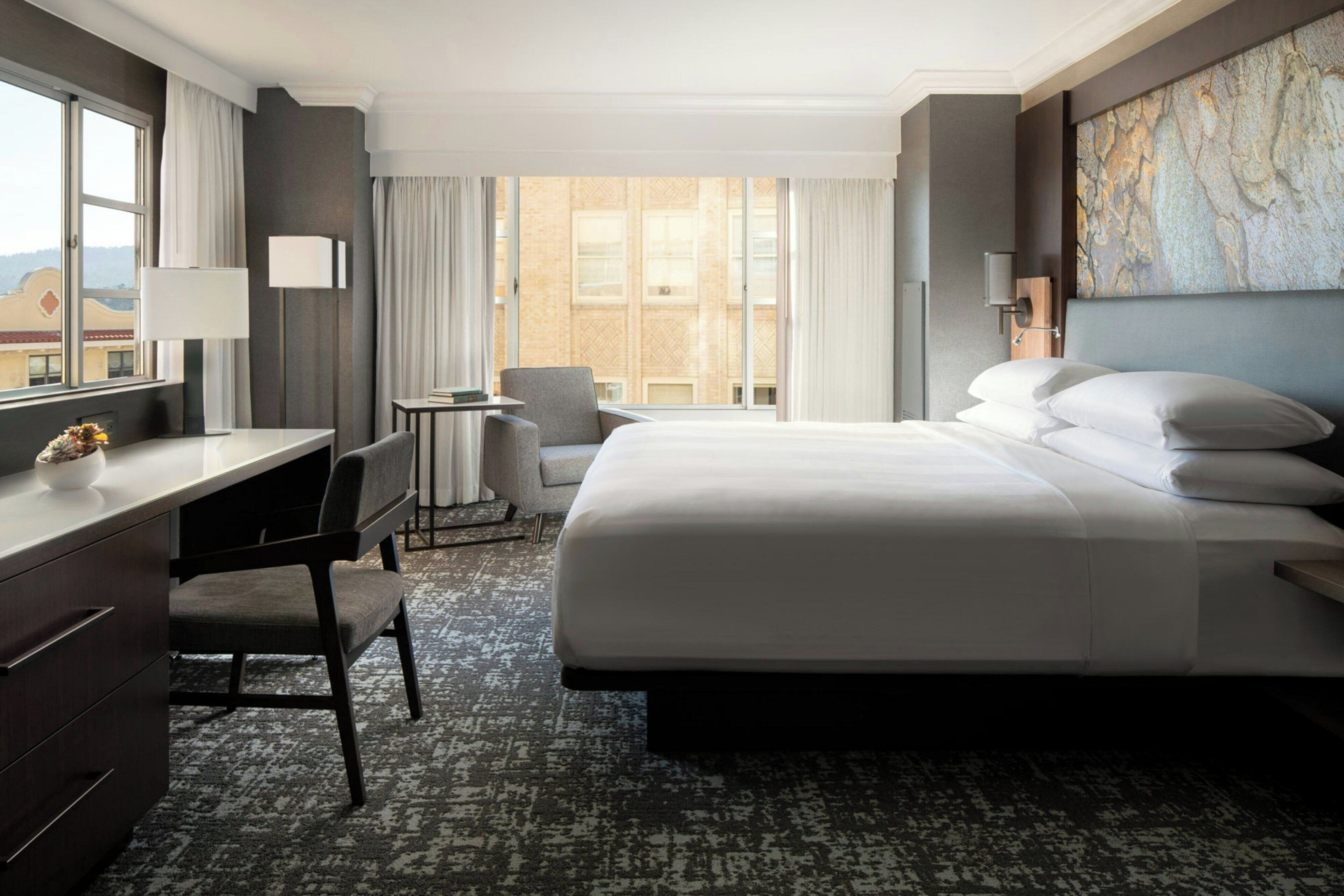 A hotel room with a bed, desk, chair, and a view of the city.