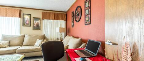 Una sala de estar con un sofá, una mesa roja con una computadora portátil, una silla y objetos decorativos en la pared.