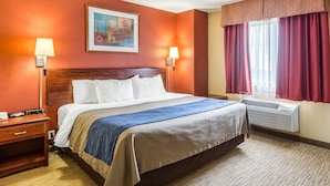 A hotel room with a bed, two bedside lamps, a painting on the wall, a window with curtains, and an air conditioning unit.