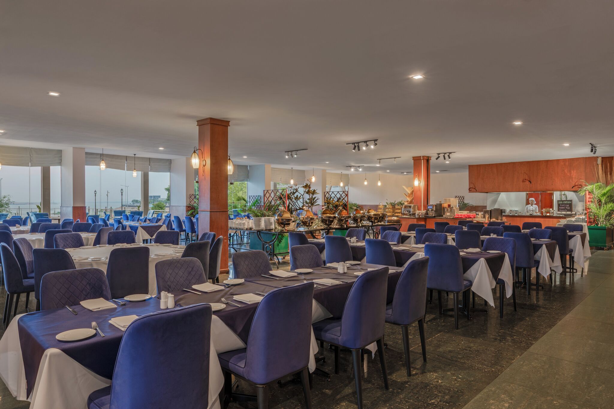 A spacious dining area with tables and chairs, a kitchen area, and large windows.
