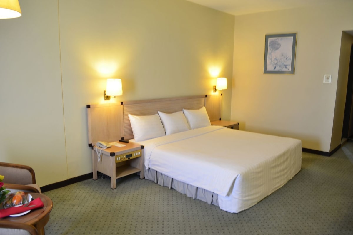 Habitación de hotel con una cama grande, lámparas de noche, una mesita de noche, una silla y una mesa de comedor con un mantel individual rojo.
