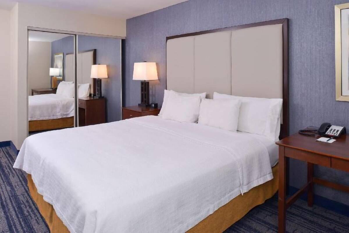 Ein Hotelzimmer mit einem großen Bett, einem Nachttisch mit Lampe, einem Spiegel und einem Telefon.