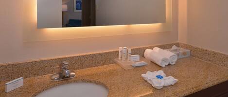 A hotel bathroom with a granite countertop sink, a mirror, and toiletries.