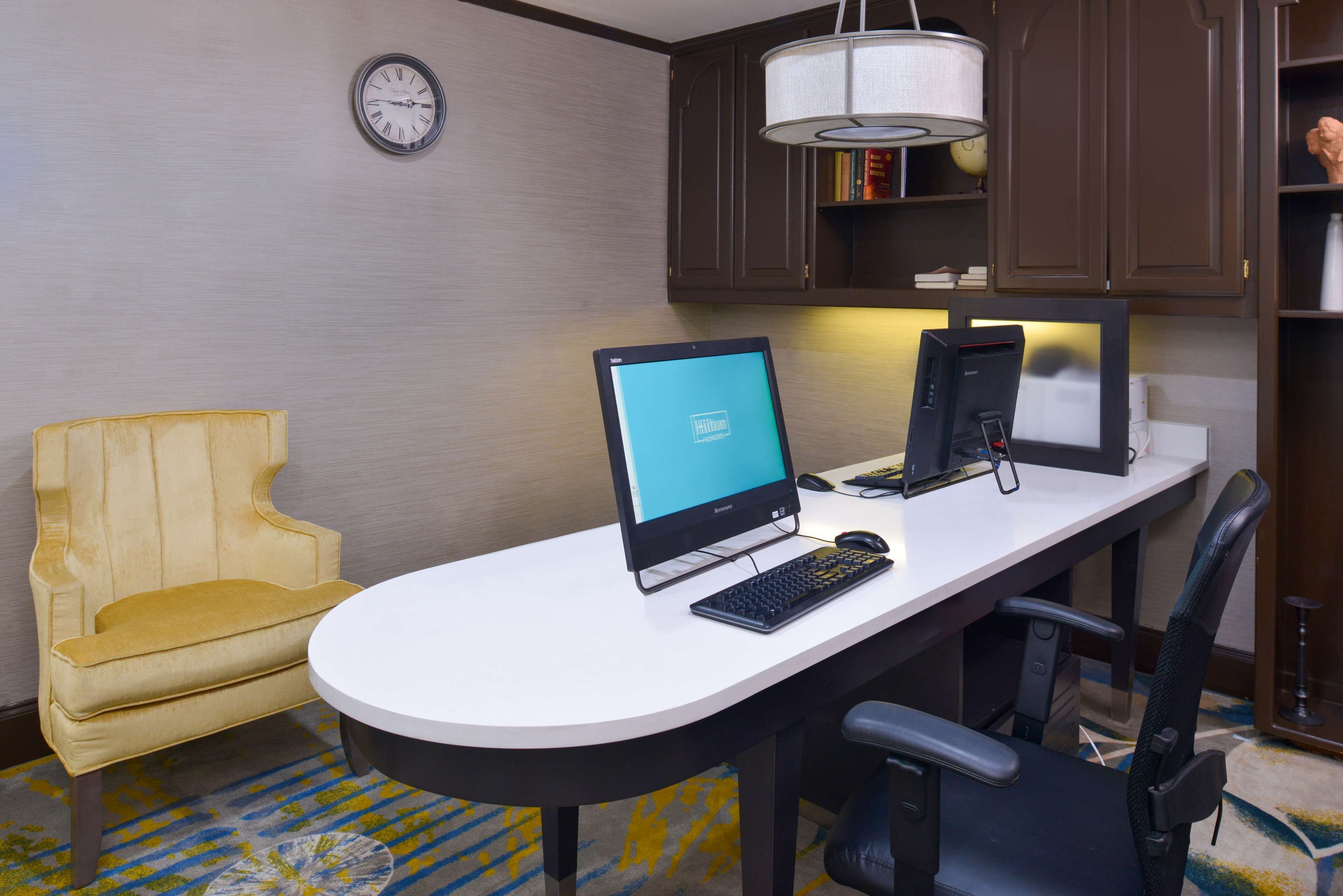 Een hotelkamer met een bureau, twee computers, een gele fauteuil, een klok en een boekenplank.