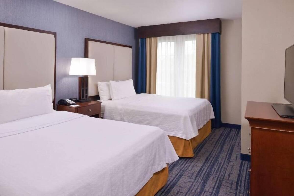 Ein Hotelzimmer mit zwei Betten, einem Nachttisch mit Lampe, einem Schreibtisch mit Laptop und einem großen Fenster mit Vorhängen.