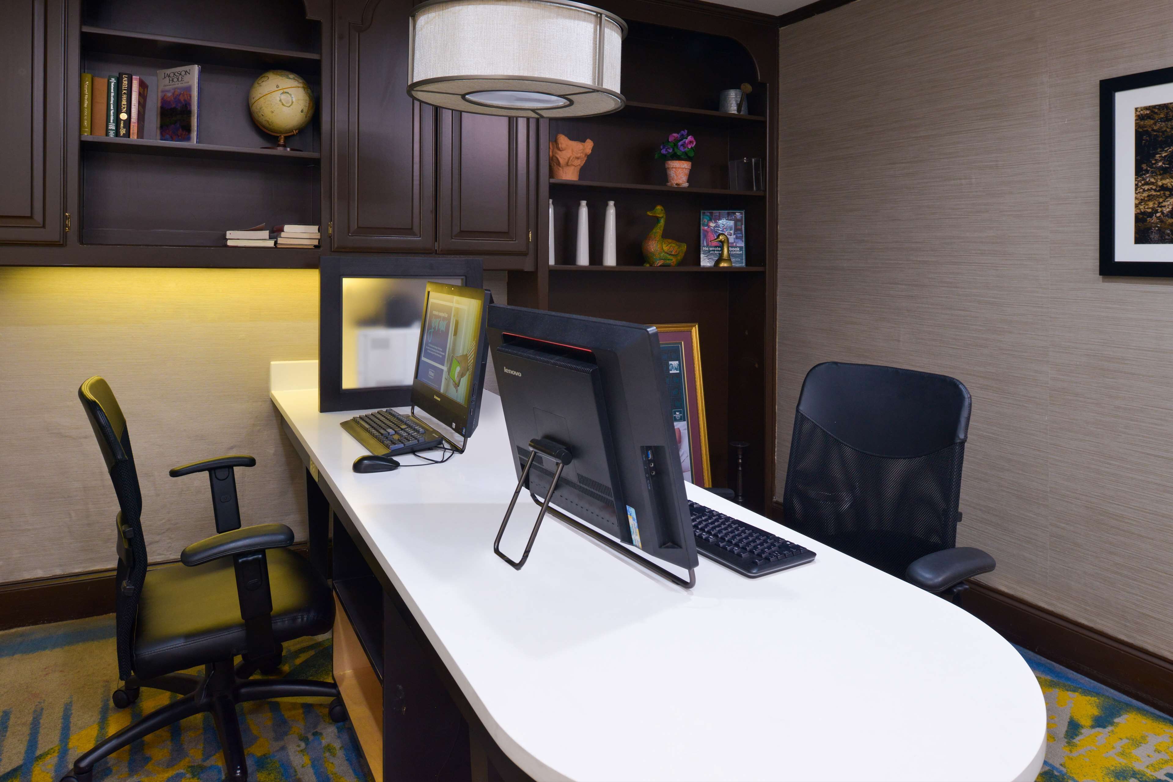 A hotel office with a desk, computer, and office chair. There are shelves with books and decorative items.
