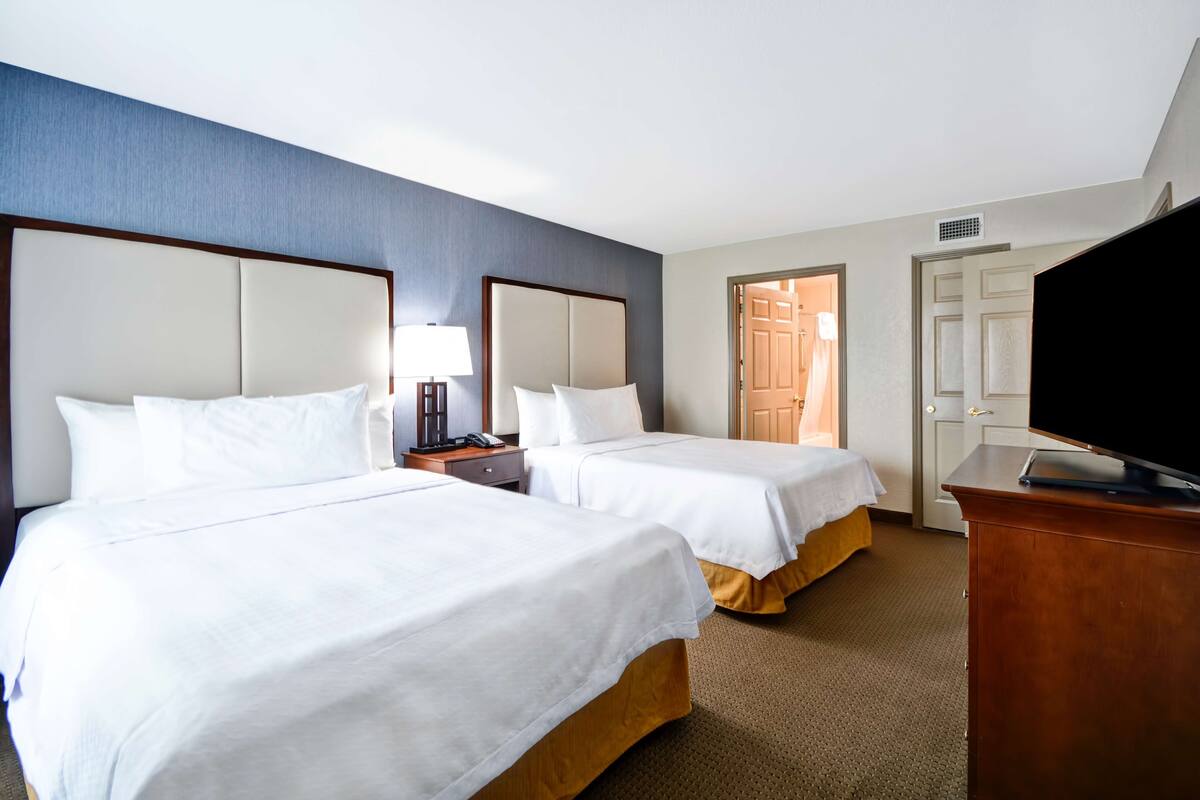 Ein Hotelzimmer mit zwei Betten, einem Fernseher, einem Schreibtisch und einer Tür zu einem weiteren Raum.
