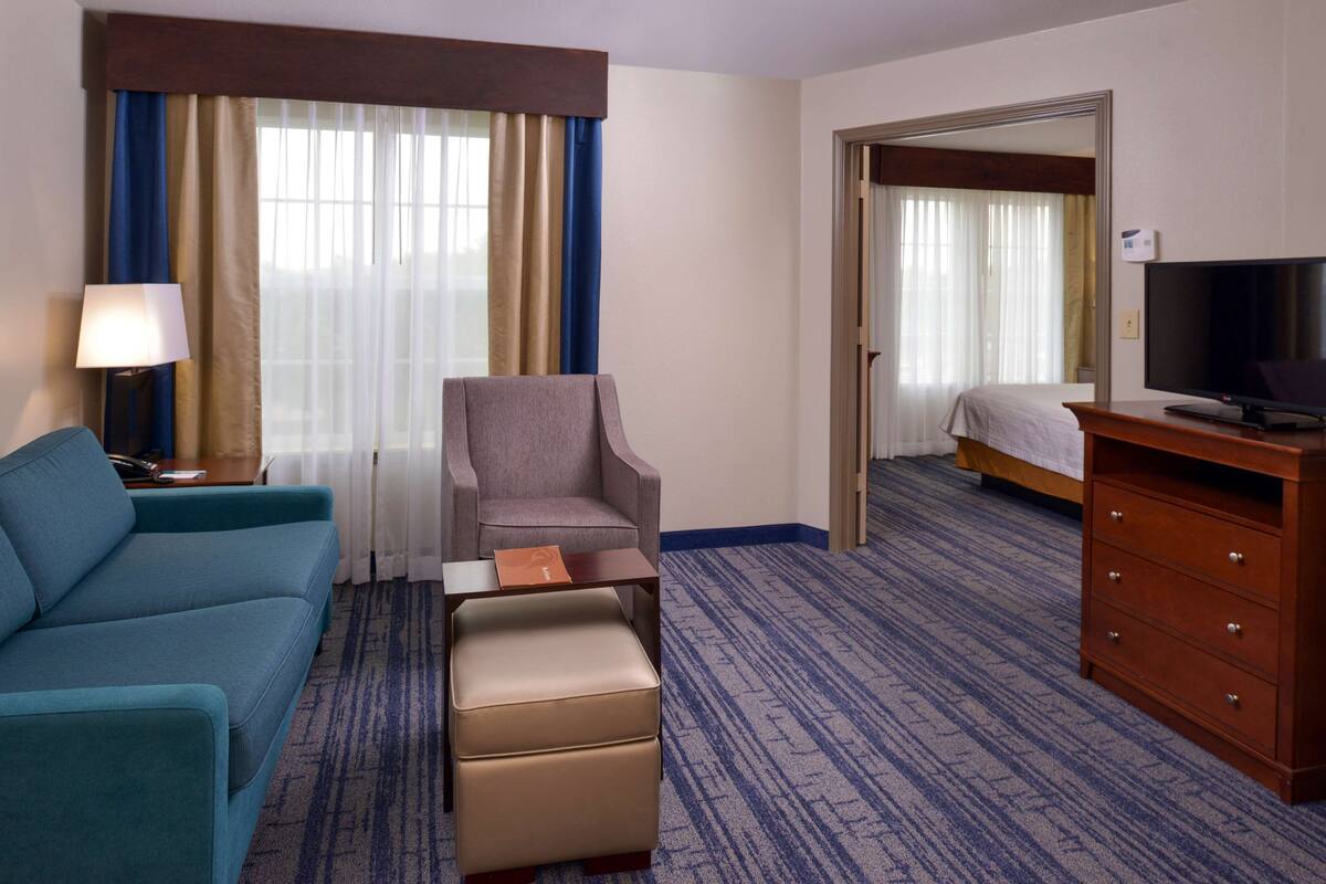 Ein Hotelzimmer mit einem blauen Sofa, einem kleinen beigen Hocker, einem Nachttisch aus Holz mit einer Lampe, einem Fernseher auf einem Holzgestell und einem großen Spiegel an der Wand.