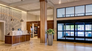 A hotel lobby with a reception desk, a large potted plant, and a view through the glass doors.