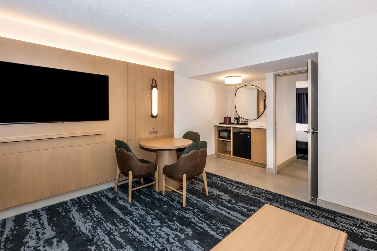 Habitación de hotel con televisión de pantalla plana, una mesa de comedor redonda con sillas, una pequeña cocina y un espejo en la pared.