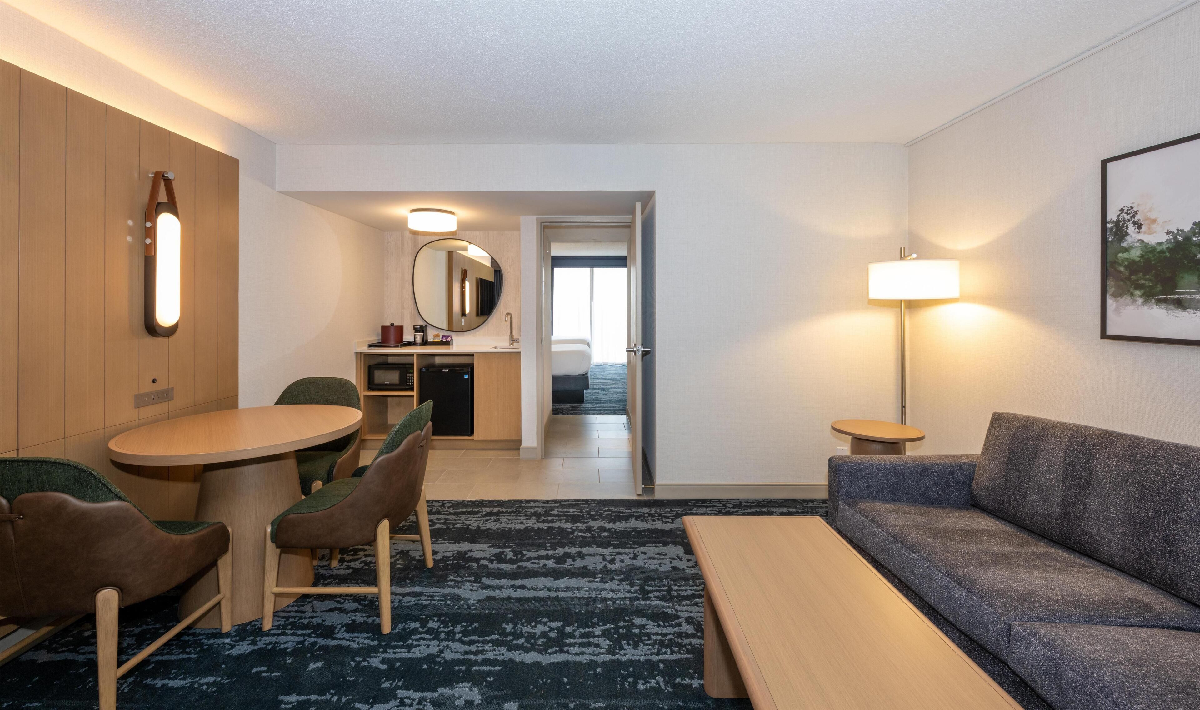 Quarto de hotel com mesa de jantar, cadeiras, sofá, luminária, micro-ondas, geladeira, espelho e porta que dá para um quarto.