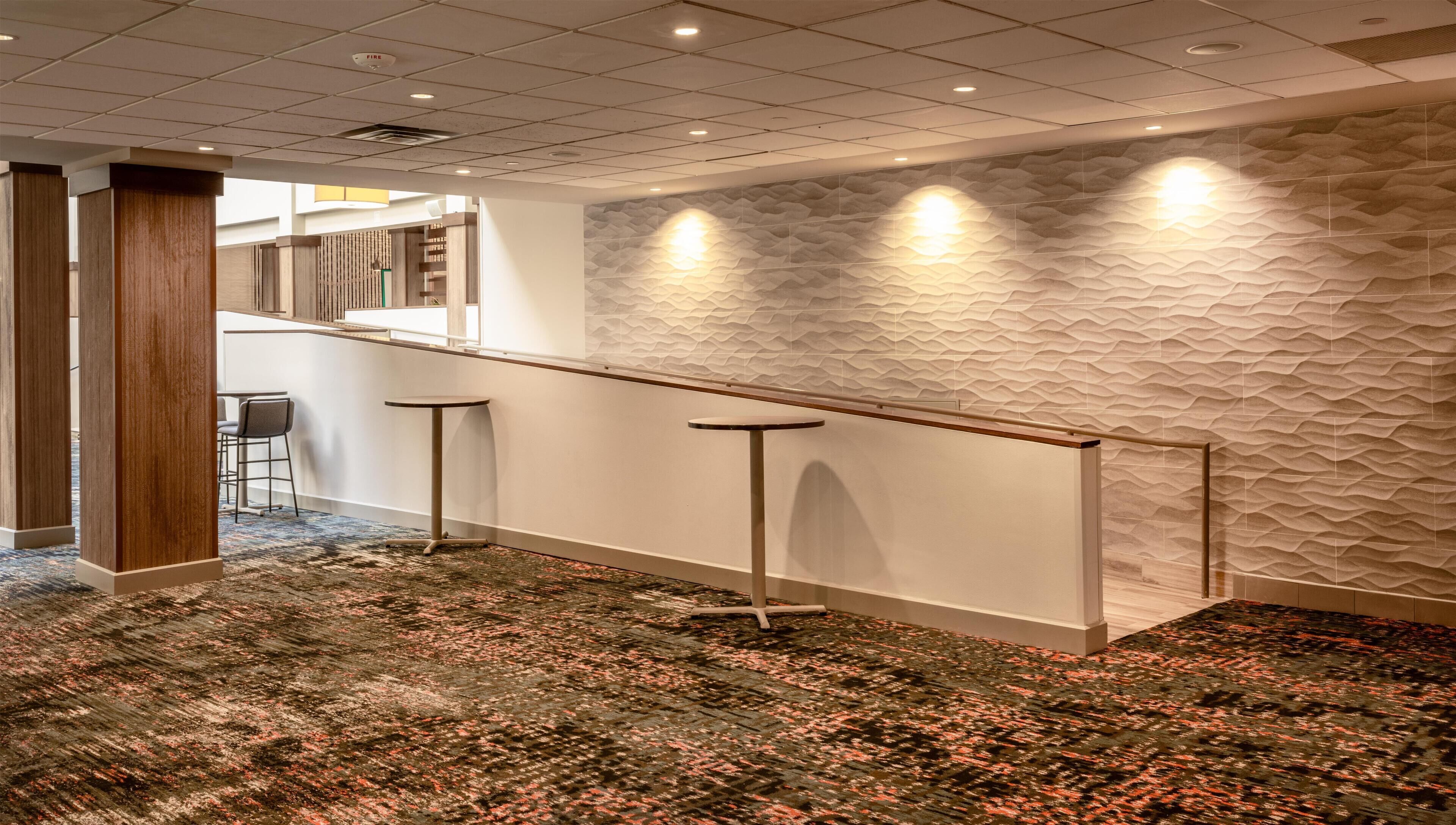 A modern hotel lobby with a high ceiling, a patterned wall, and a bar area with stools.