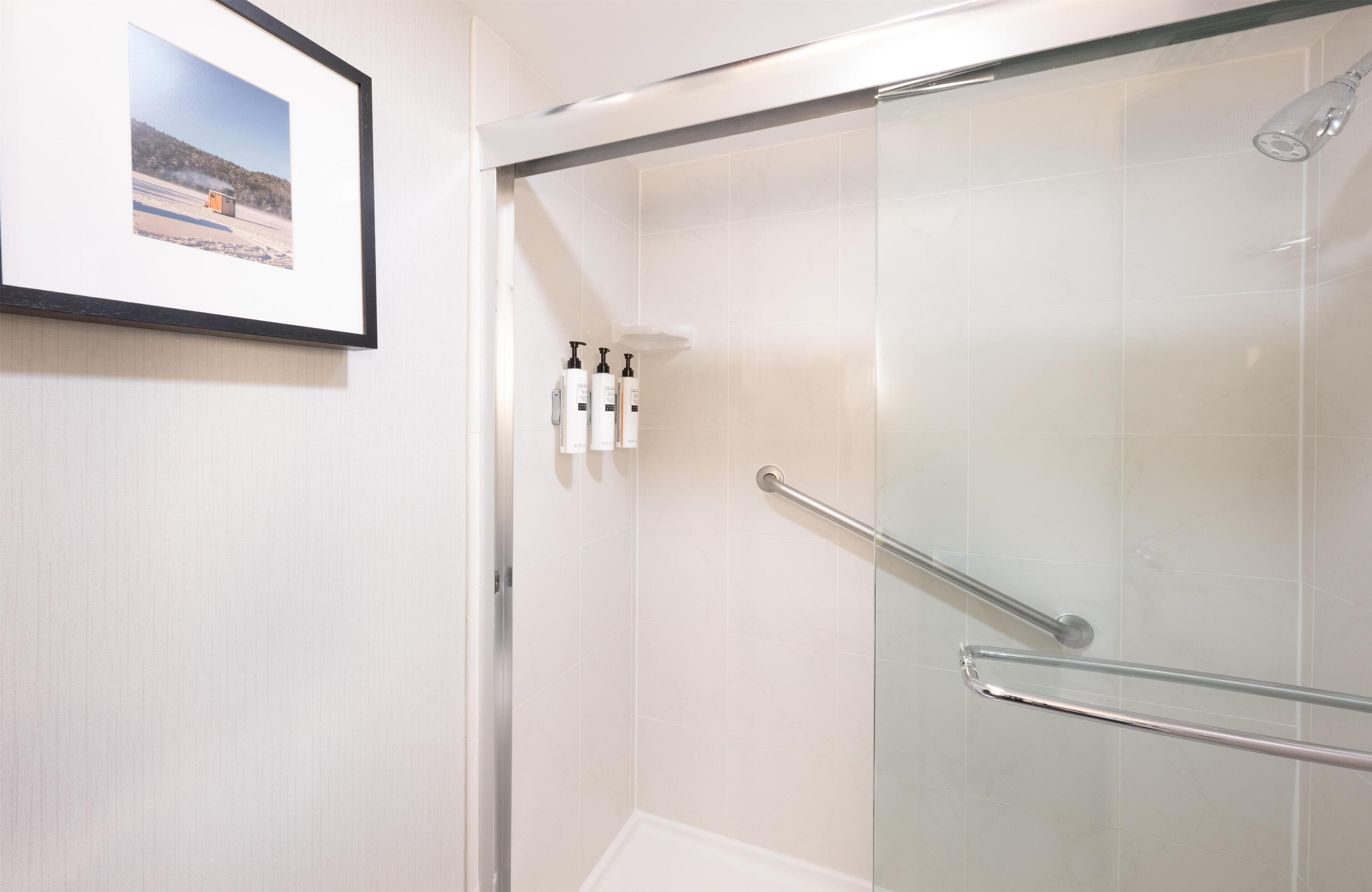 A shower with a glass door, a framed picture on the wall, and toiletries on a shelf.
