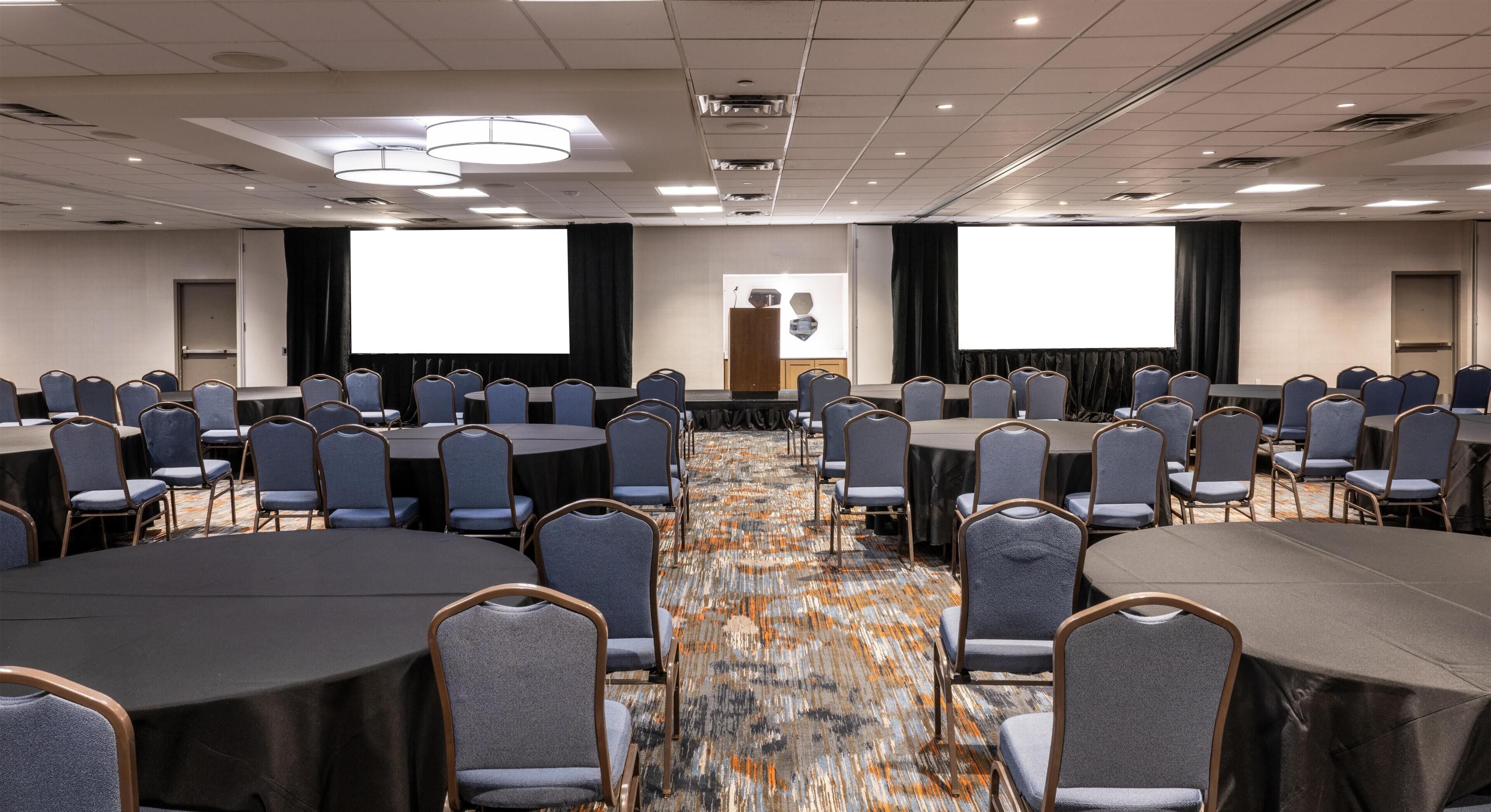 A conference room with round tables, chairs, a carpeted floor, and a projector screen.