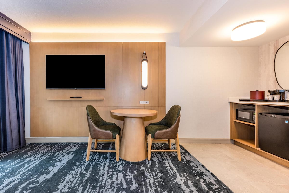 Habitación de hotel con televisor de pantalla plana montado en la pared, un mueble de madera debajo, una mesa redonda con dos sillas, microondas y refrigerador.