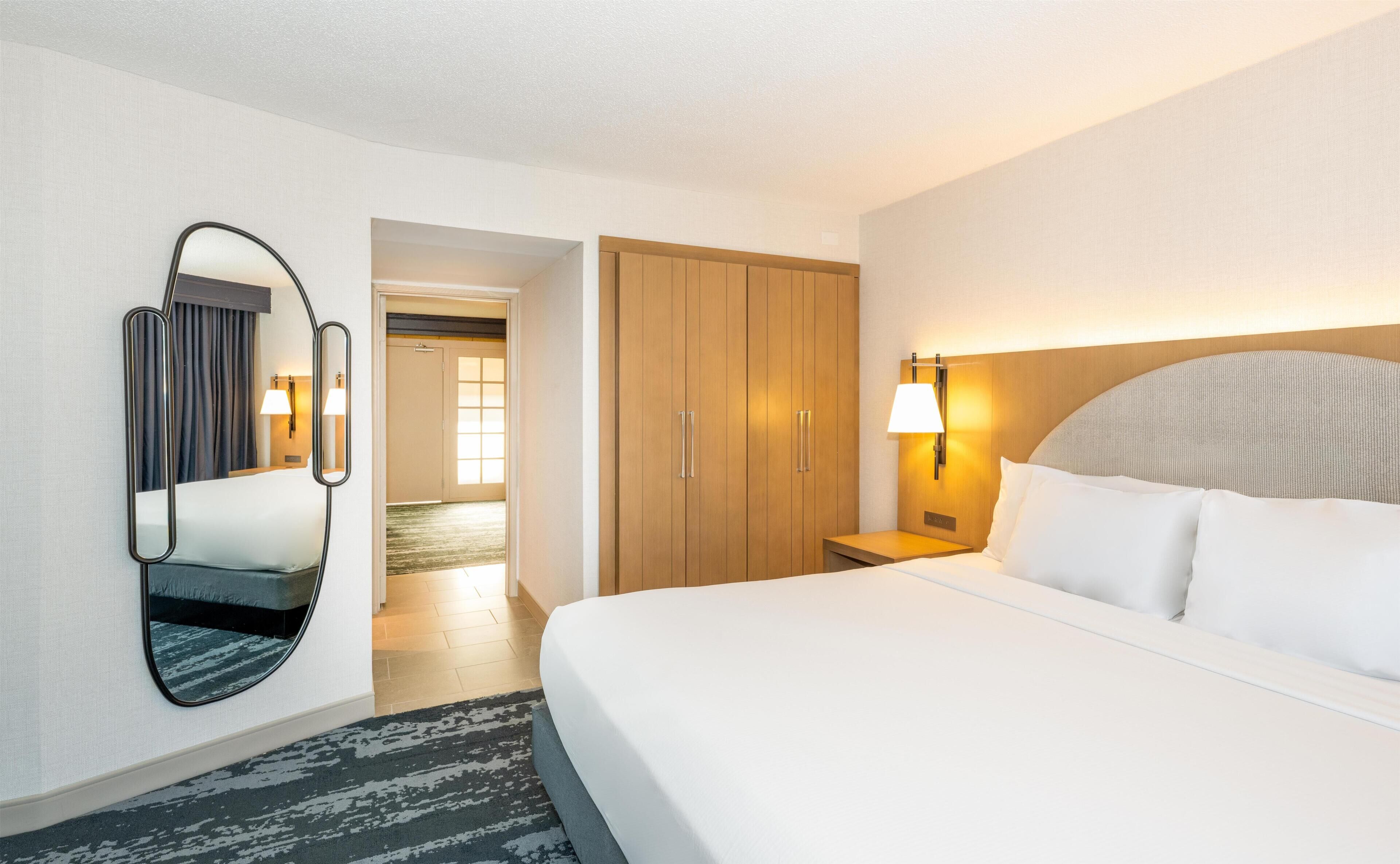 A hotel room with a bed, a nightstand, a lamp, a wardrobe, and a mirror.