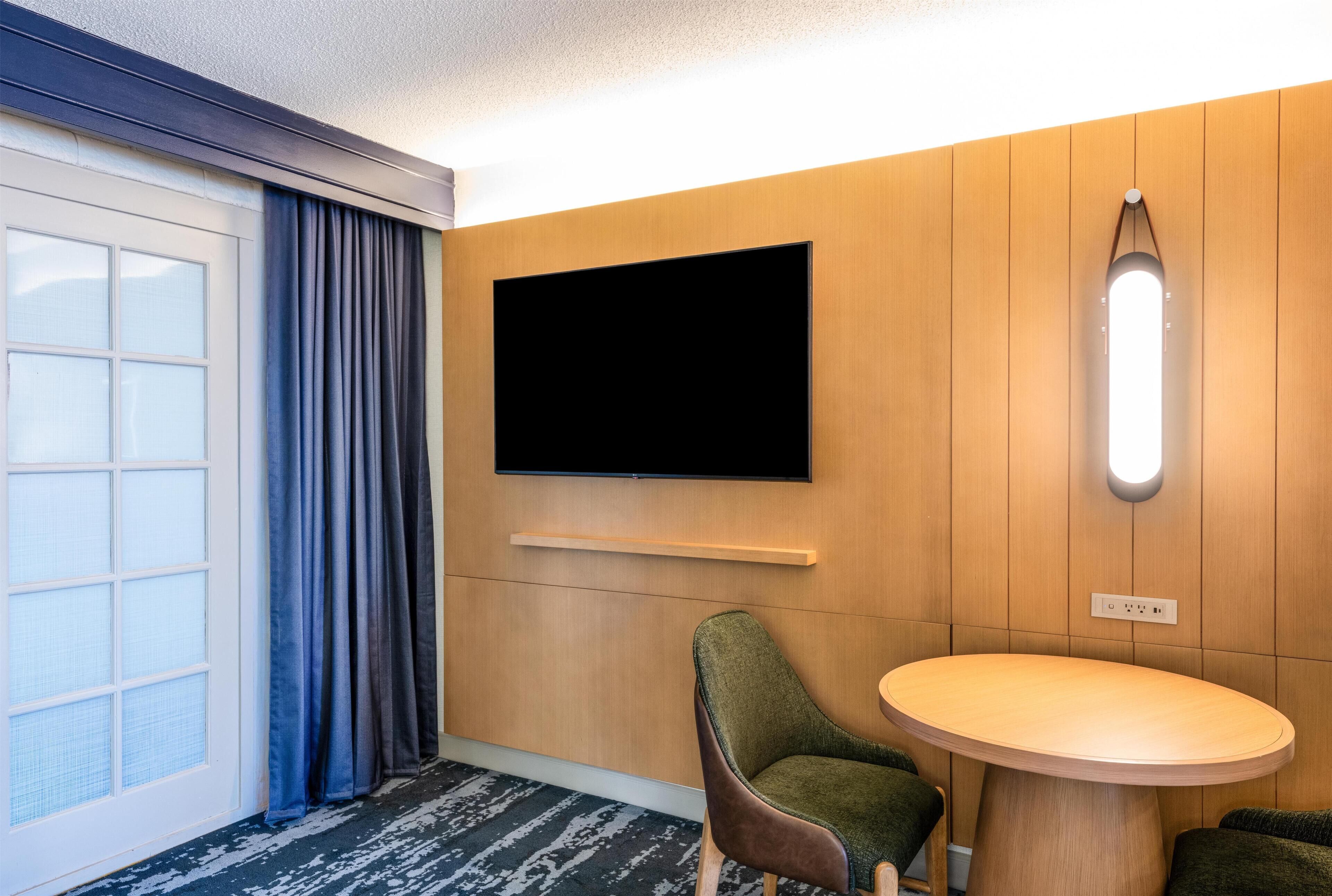A hotel room with a large window, a flat-screen TV mounted on the wall, a round wooden table, and a green upholstered chair.