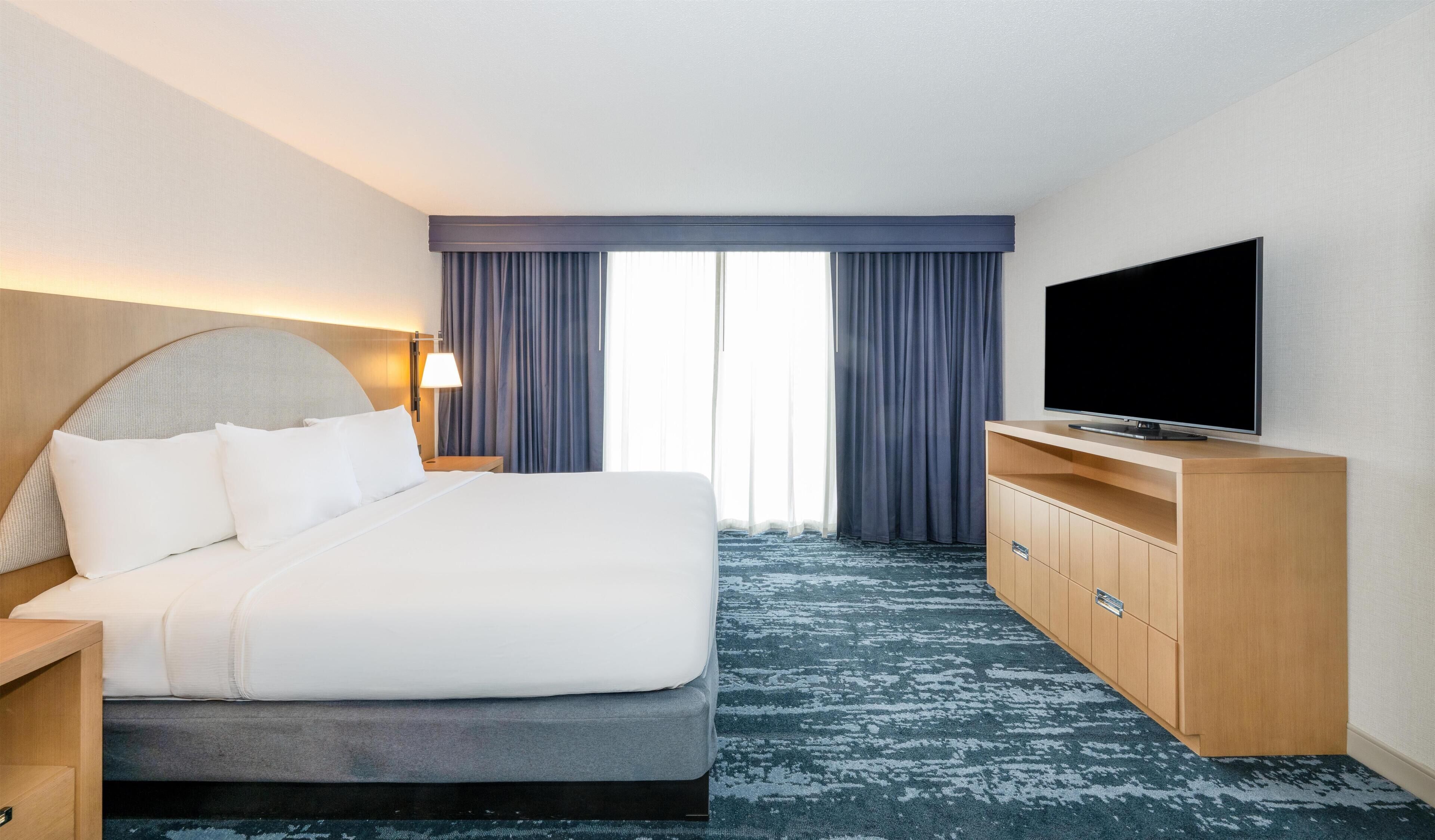 A hotel room with a bed, a TV, a desk, and a large window with curtains.