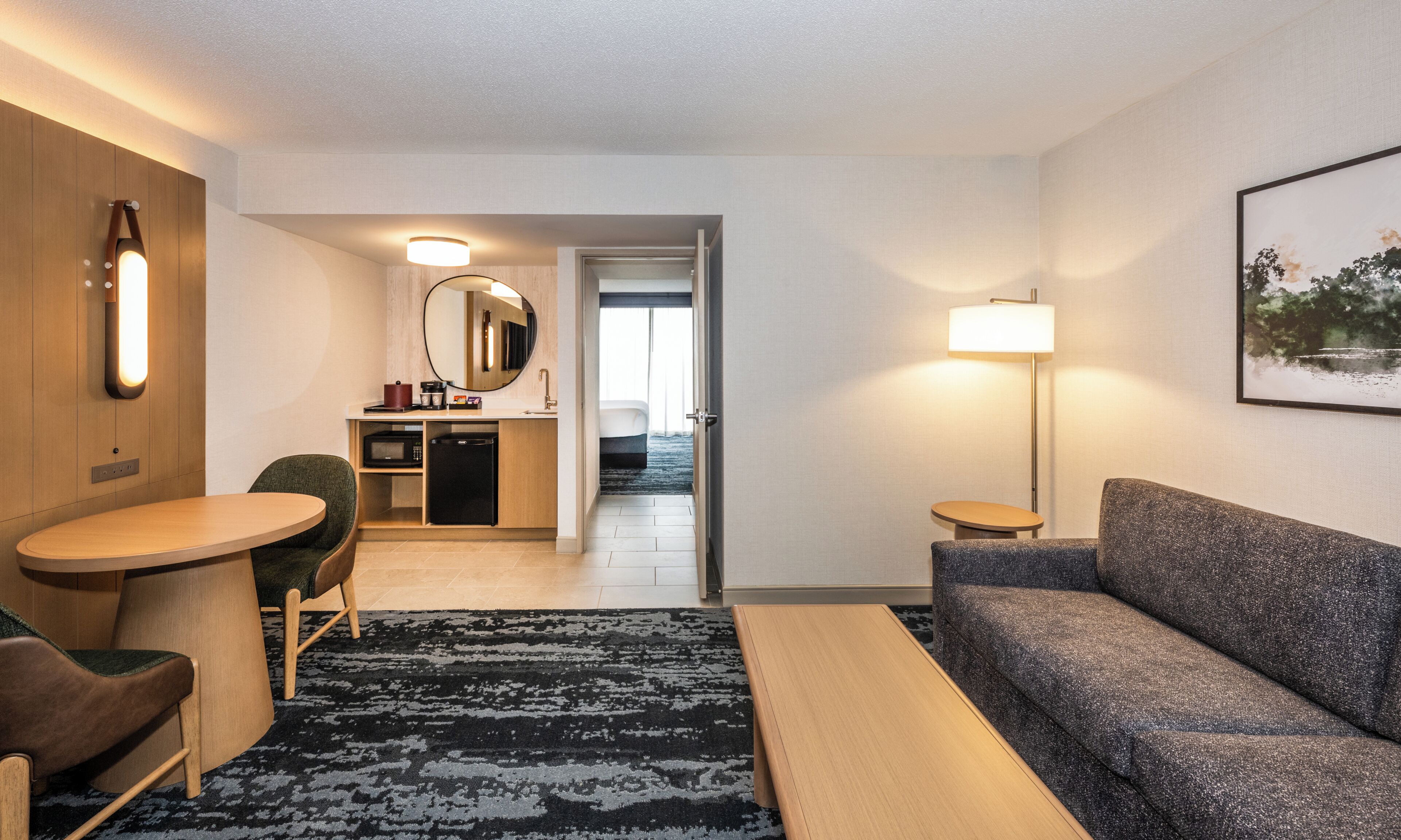 A hotel room with a sofa, a small table, a chair, a desk with a mirror, a lamp, and a painting on the wall.