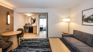A hotel room with a sofa, a small table, a chair, a desk with a mirror, a lamp, and a painting on the wall.