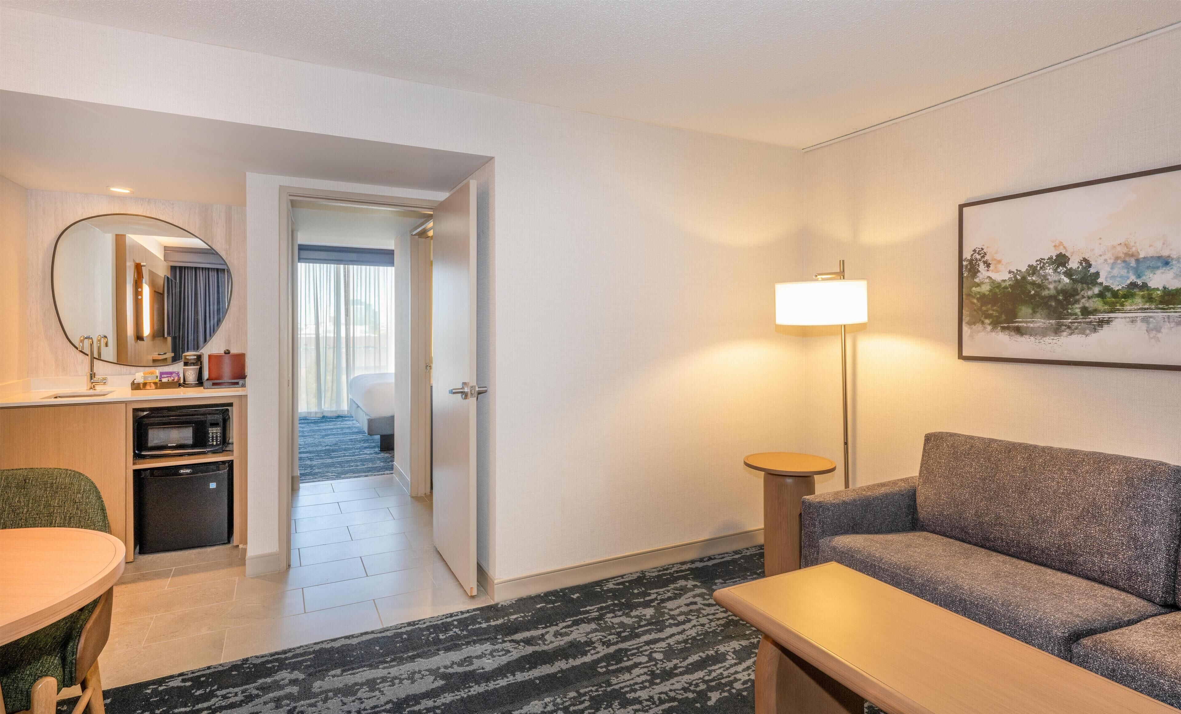 A hotel room with a sofa, a dining table, a microwave, a coffee maker, a lamp, and a painting on the wall.
