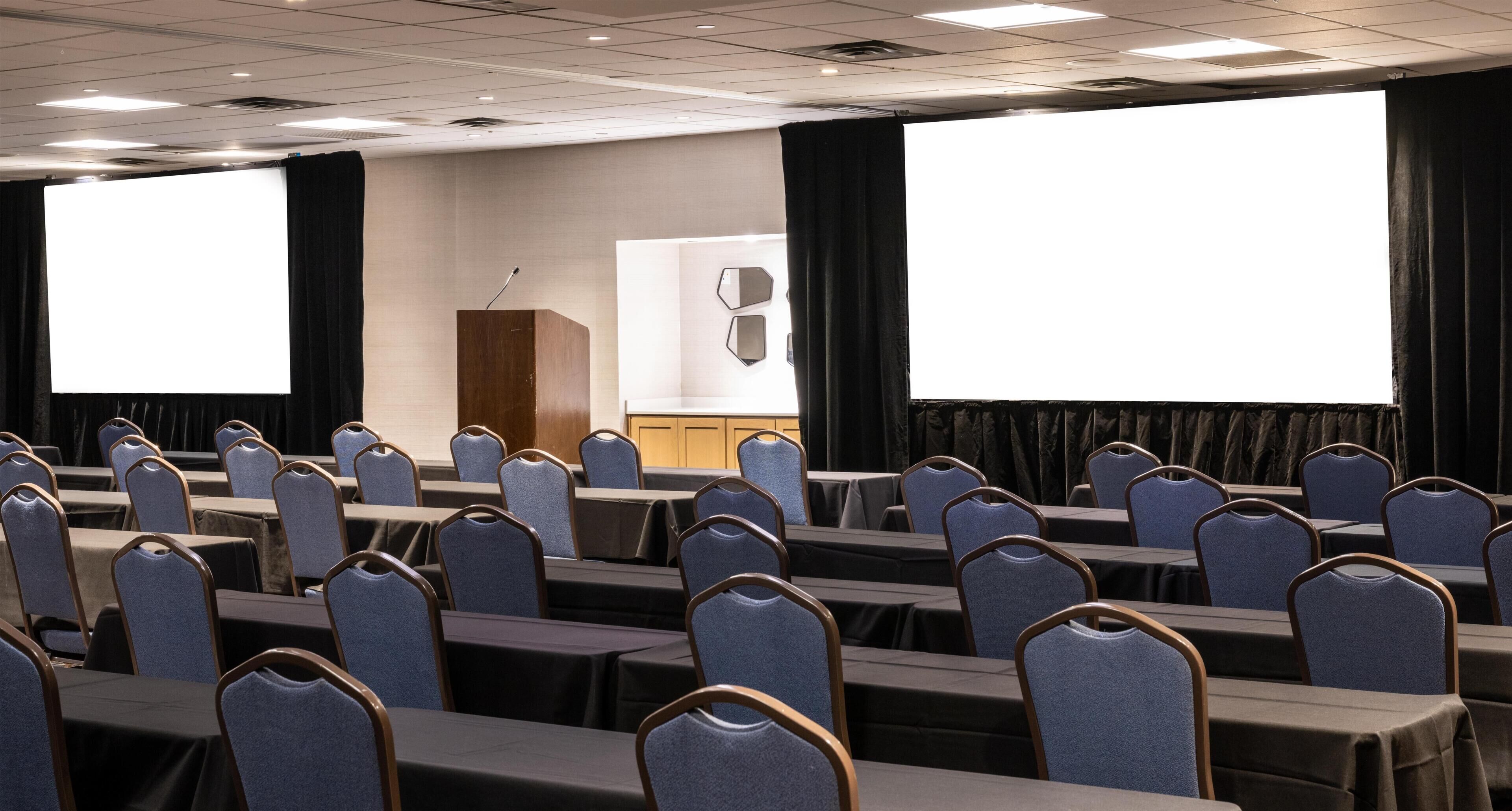 A conference room with rows of chairs, a podium, and two large screens.