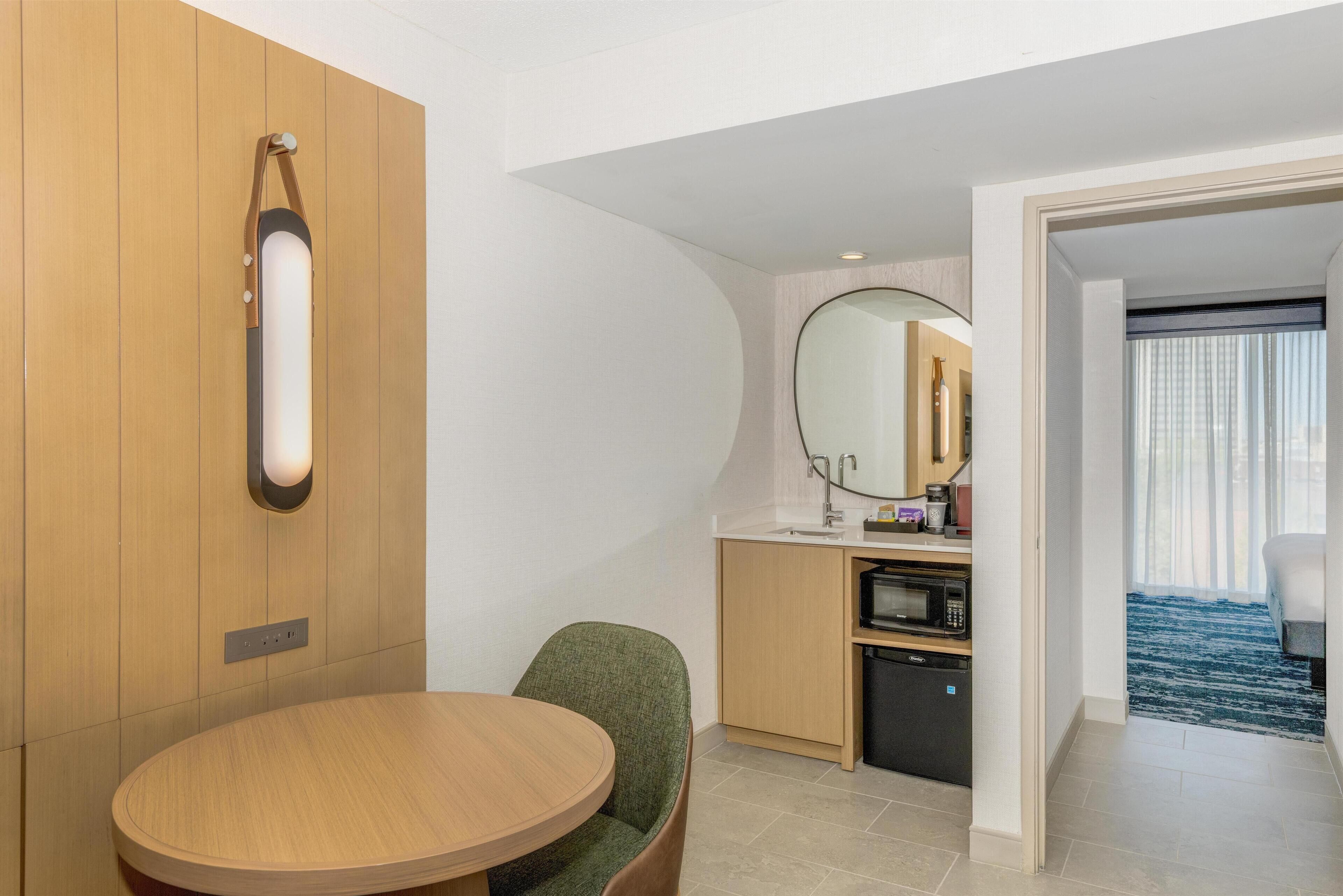 Habitación de hotel moderna con pared de madera, una mesa redonda, una silla verde, una zona de cocina con microondas y un espejo grande.