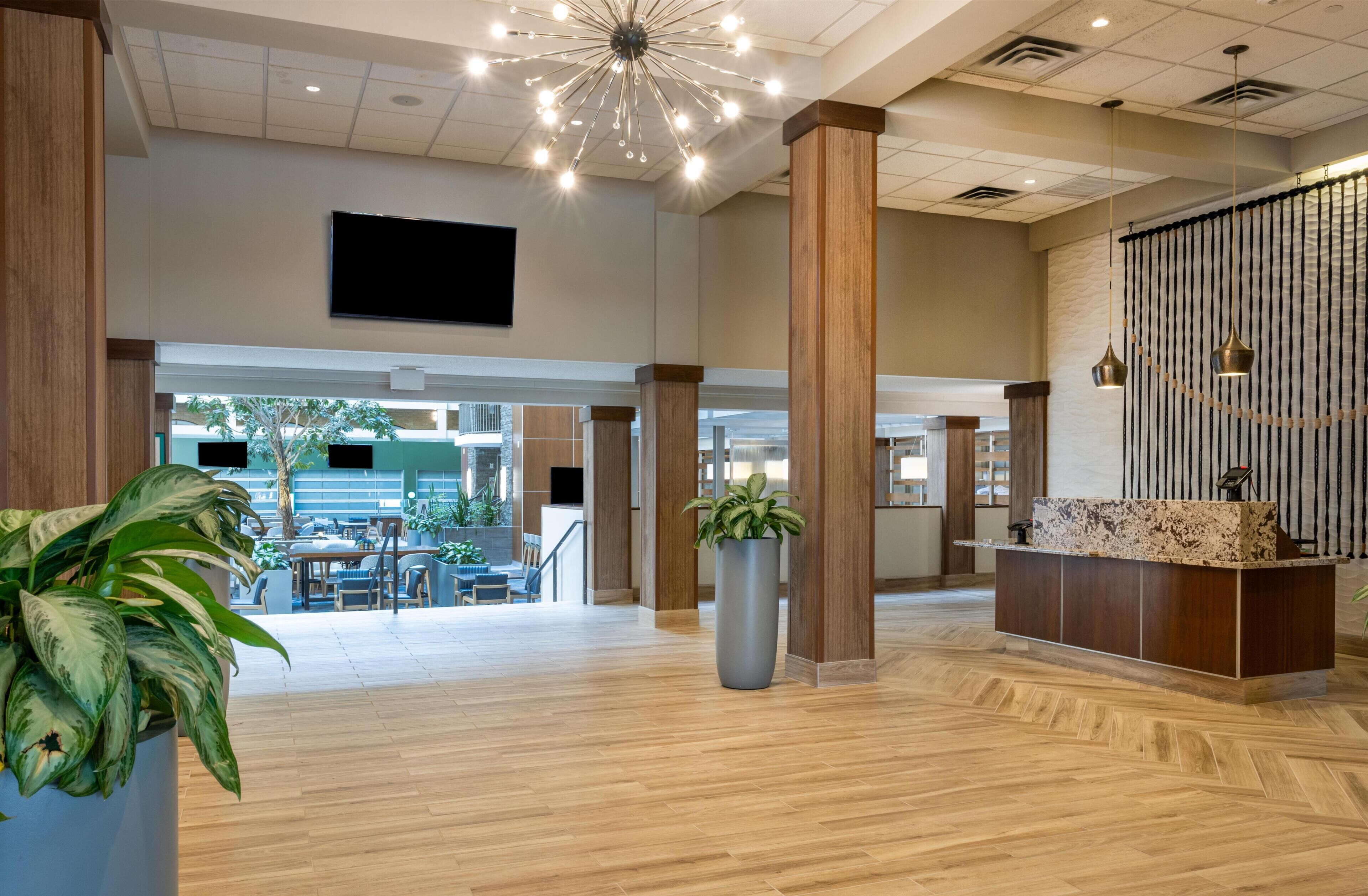 A spacious lobby with a reception desk, wooden pillars, a large potted plant, and a modern chandelier.