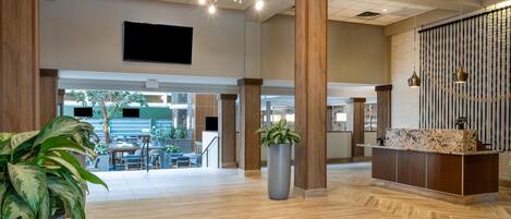 A spacious lobby with a reception desk, wooden pillars, a large potted plant, and a modern chandelier.