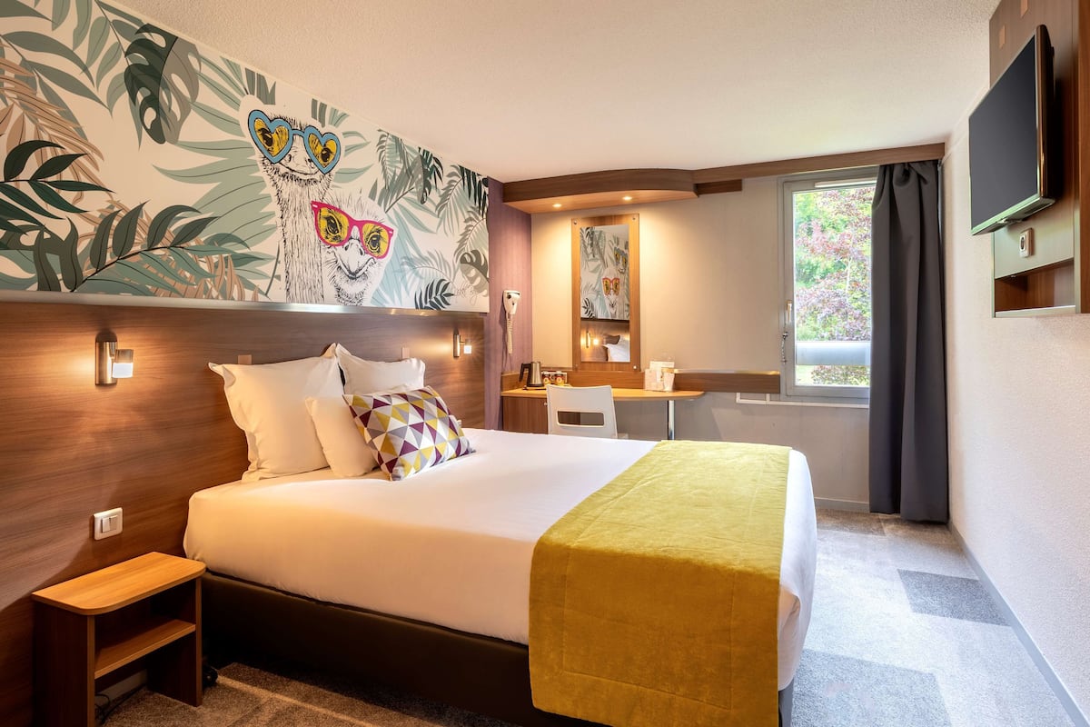 Une chambre d’hôtel avec un lit, un bureau, une chaise, une télévision et une fenêtre avec des rideaux.