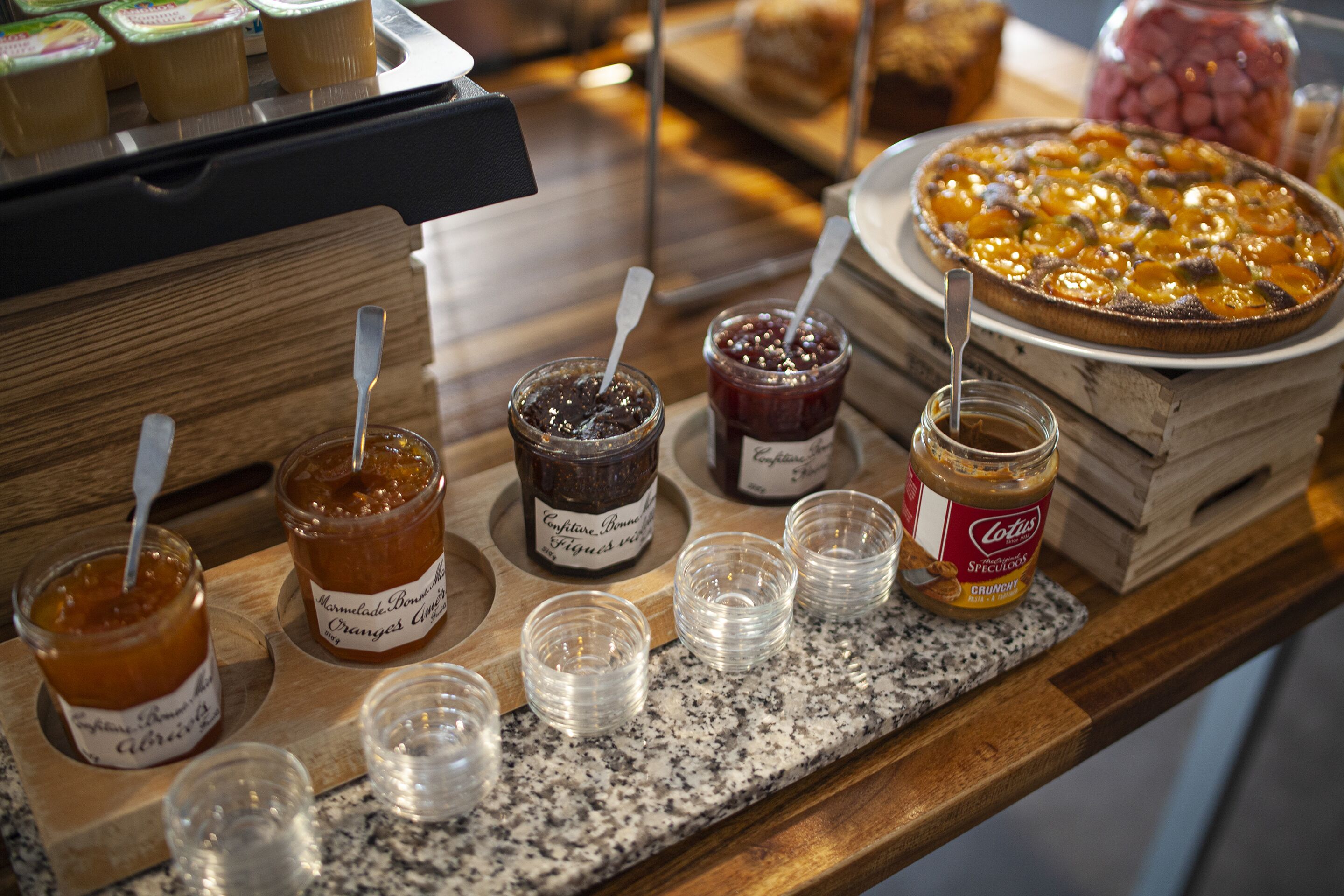 A variety of food items including jars of jam and a large pie on a wooden counter.