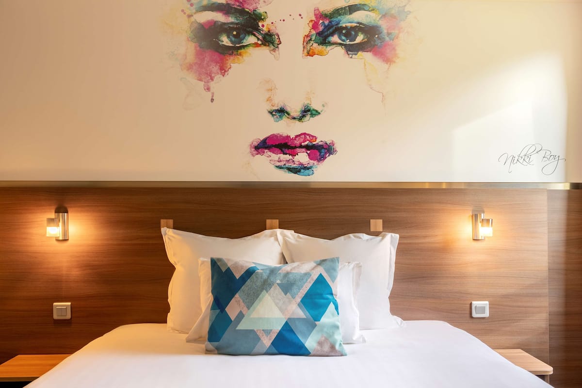 Une chambre d’hôtel avec une tête de lit en bois, deux tables de chevet et une peinture abstraite aux couleurs vives accrochée au mur.