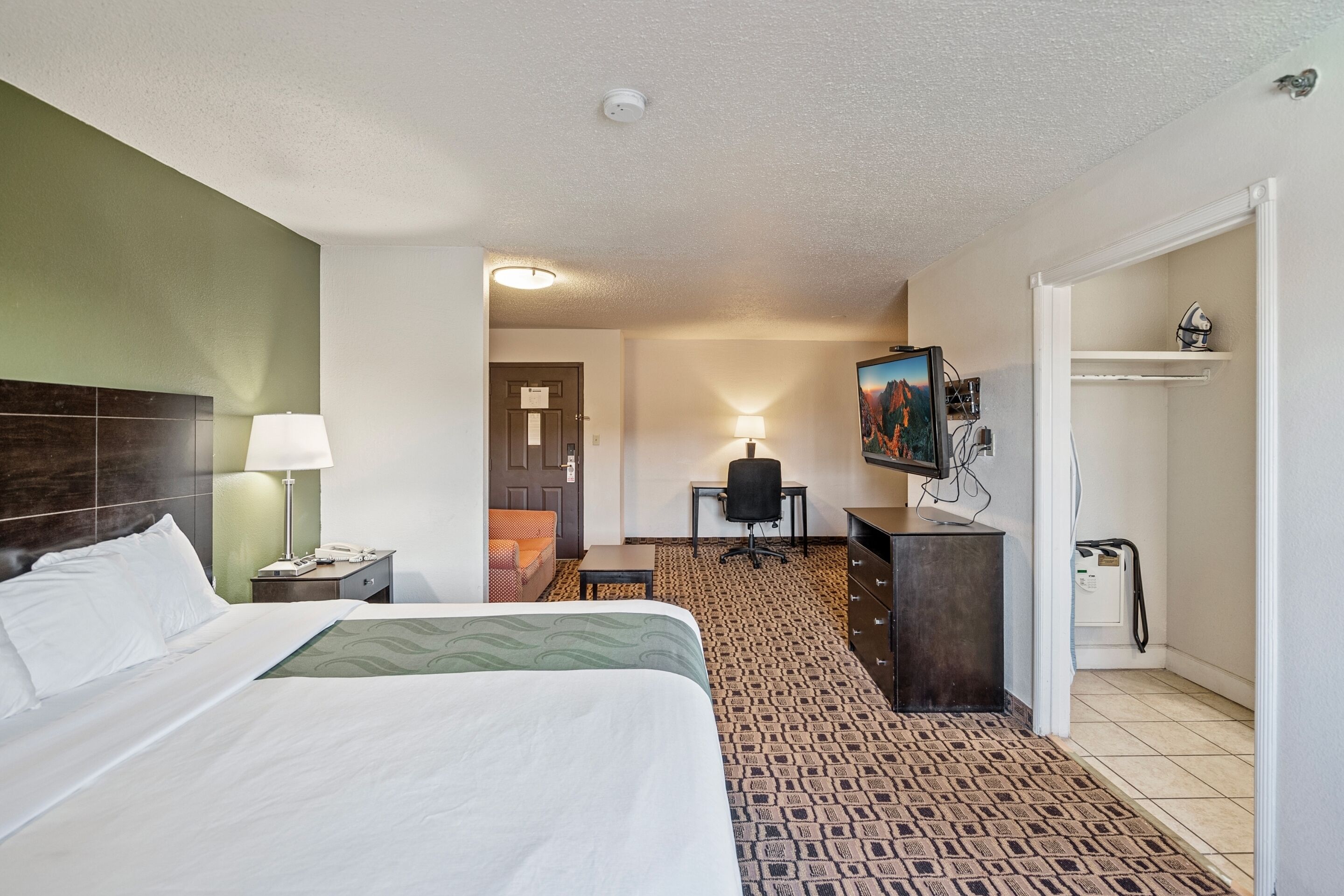 A hotel room with a bed, a desk, a chair, a television, and a coat rack.