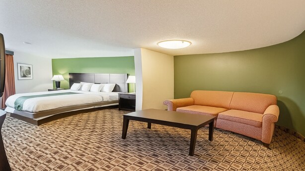 A hotel room with a bed, a sofa, a table, and a patterned carpet. - Coratel Plus Suites Wichita West Airport (Wichita)