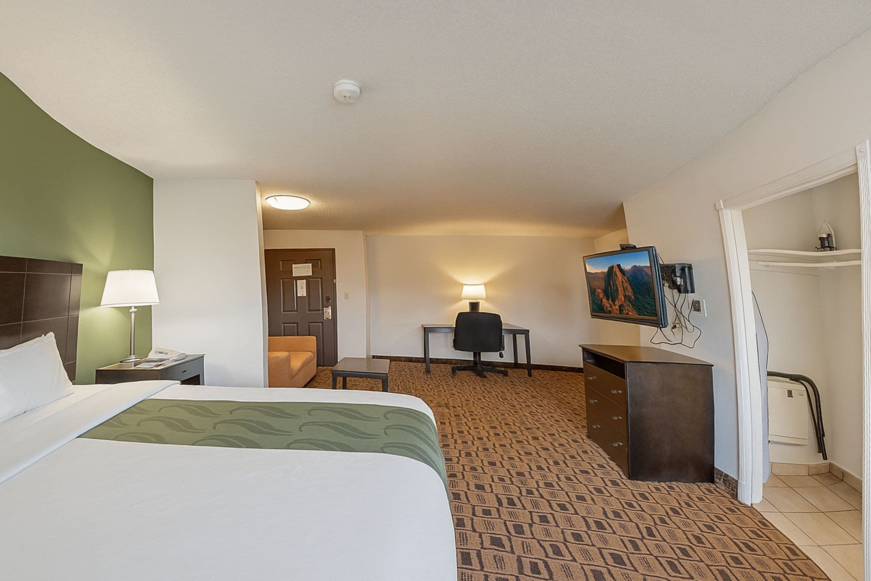 A hotel room with a bed, desk, chair, lamp, and a painting on the wall.