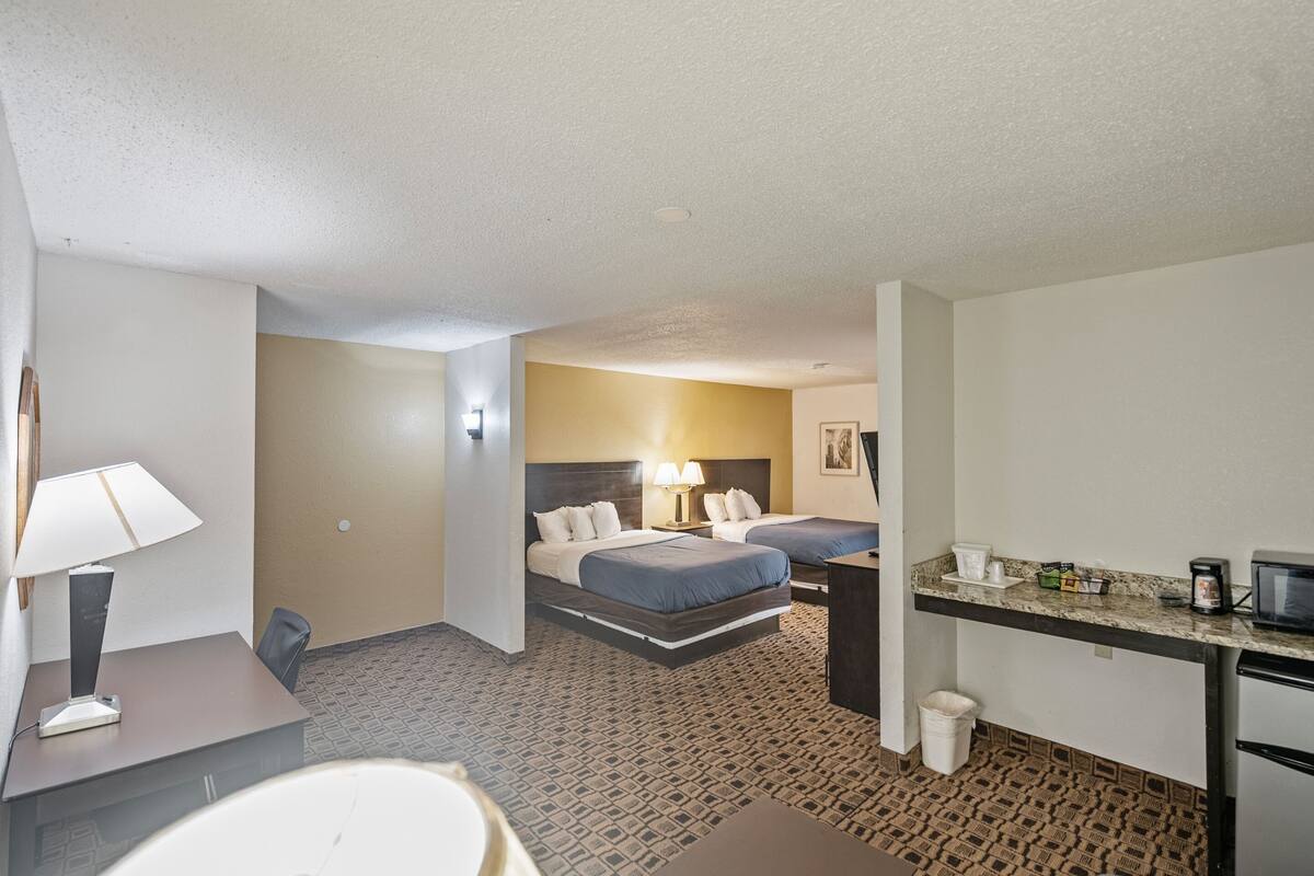 A hotel room with two beds, a desk, a chair, a small kitchenette, and a bathroom.