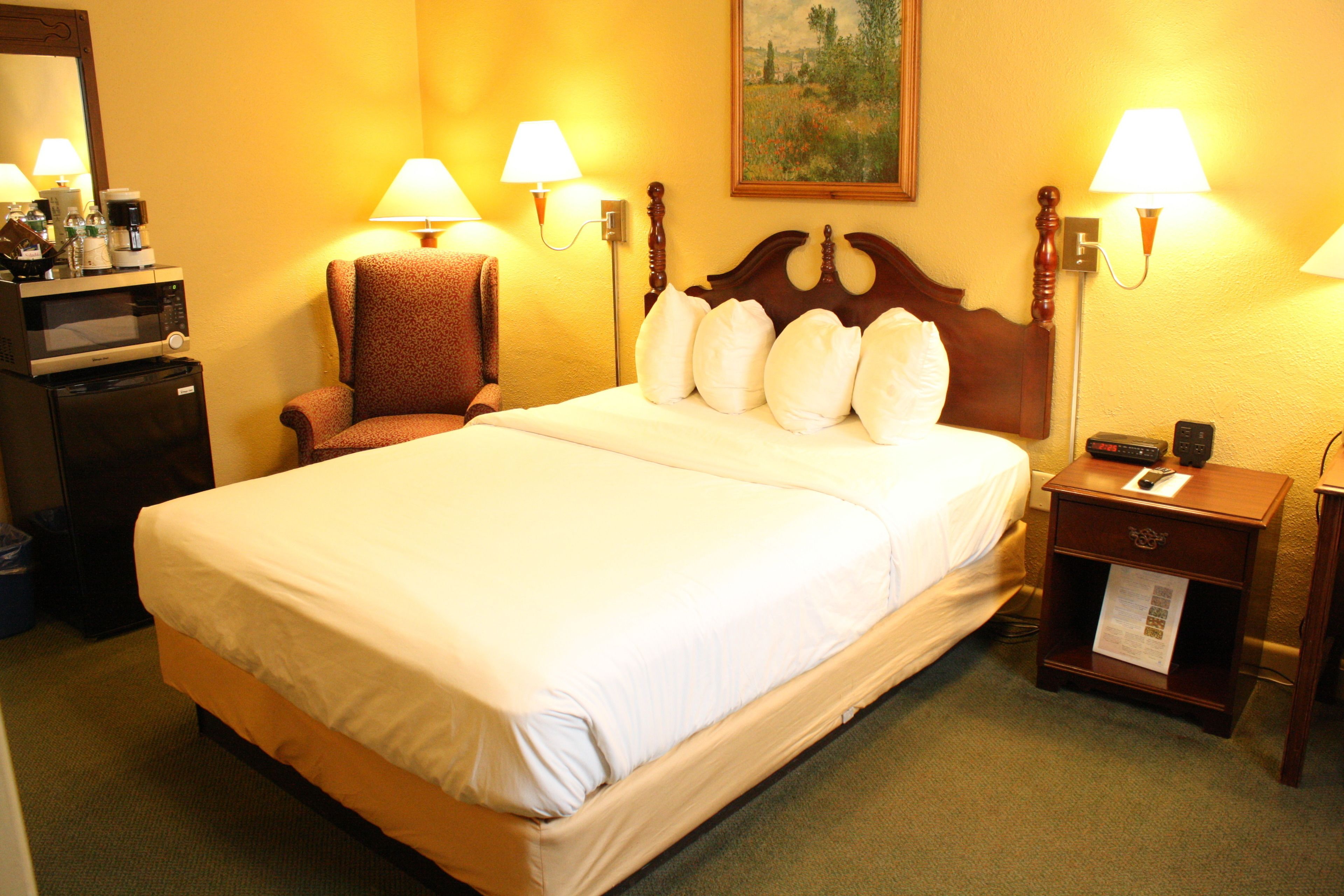 Une chambre d’hôtel avec un grand lit, deux lampes de chevet, une chaise, un bureau avec une machine à café et un tableau au mur.
