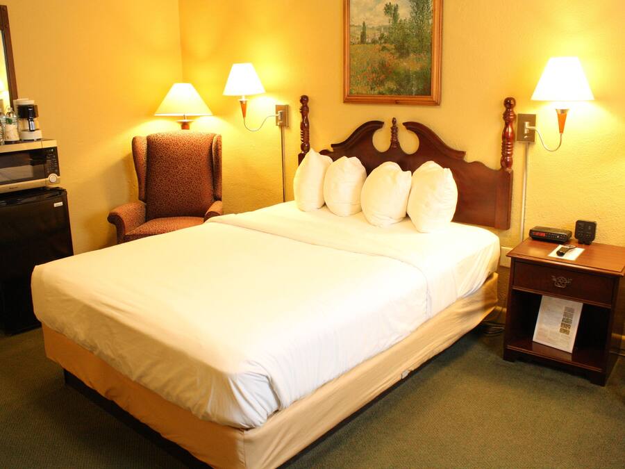 Una habitación de hotel con una cama grande, dos lámparas de noche, una silla, un escritorio con cafetera y un cuadro en la pared.