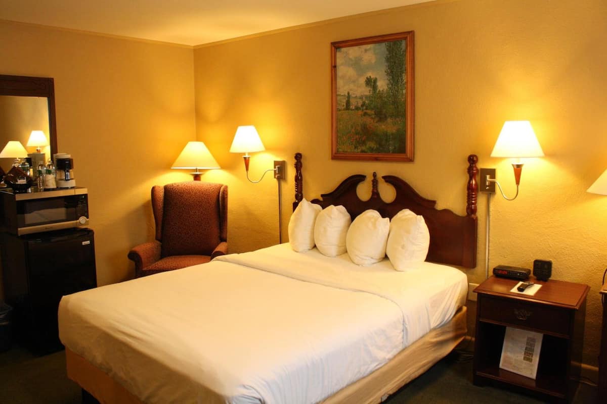 A hotel room with a large bed, two bedside lamps, a wooden headboard, a small table with a lamp, a chair, a desk with a coffee maker, and a painting on the wall.