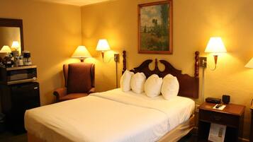 A hotel room with a large bed, two bedside lamps, a wooden headboard, a small table with a lamp, a chair, a desk with a coffee maker, and a painting on the wall.