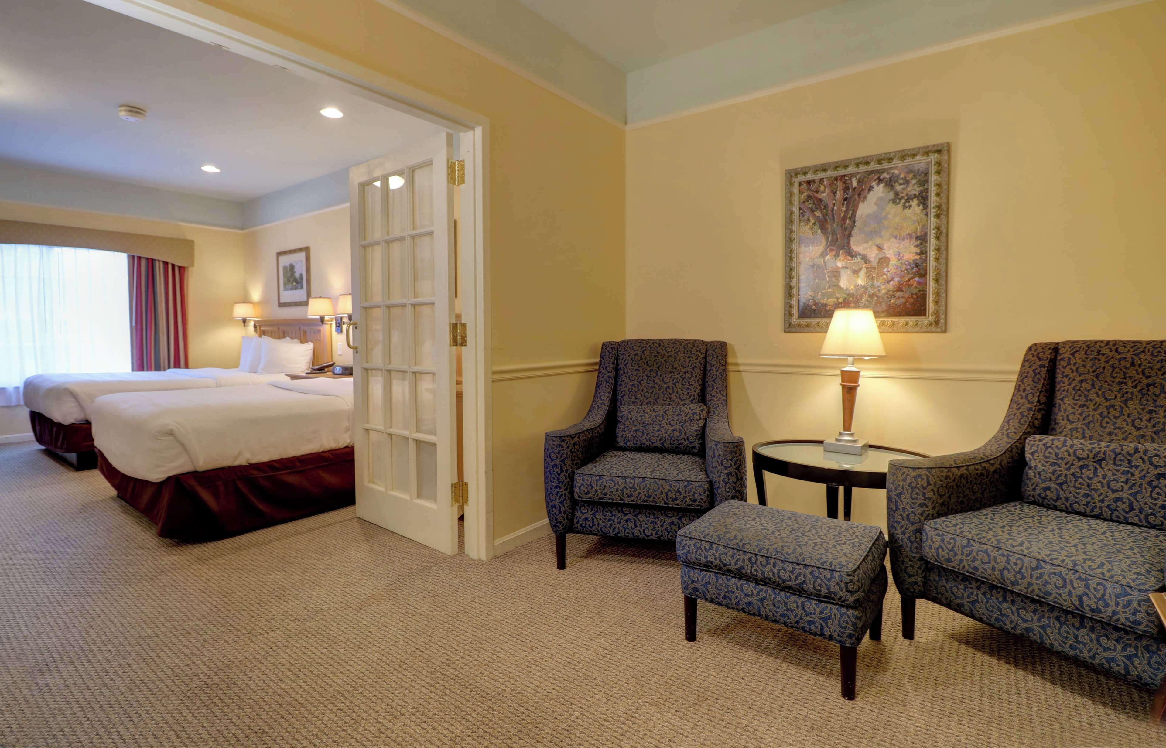 A hotel room with two beds, a sitting area with a chair and ottoman, a small table with a lamp, and a painting on the wall.