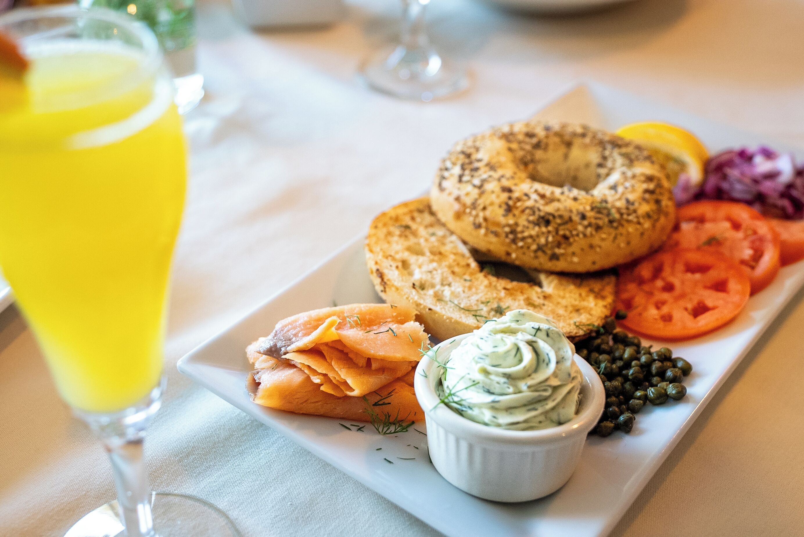 A breakfast plate with bagels, smoked salmon, tomatoes, capers, and a bowl of cream cheese dip.