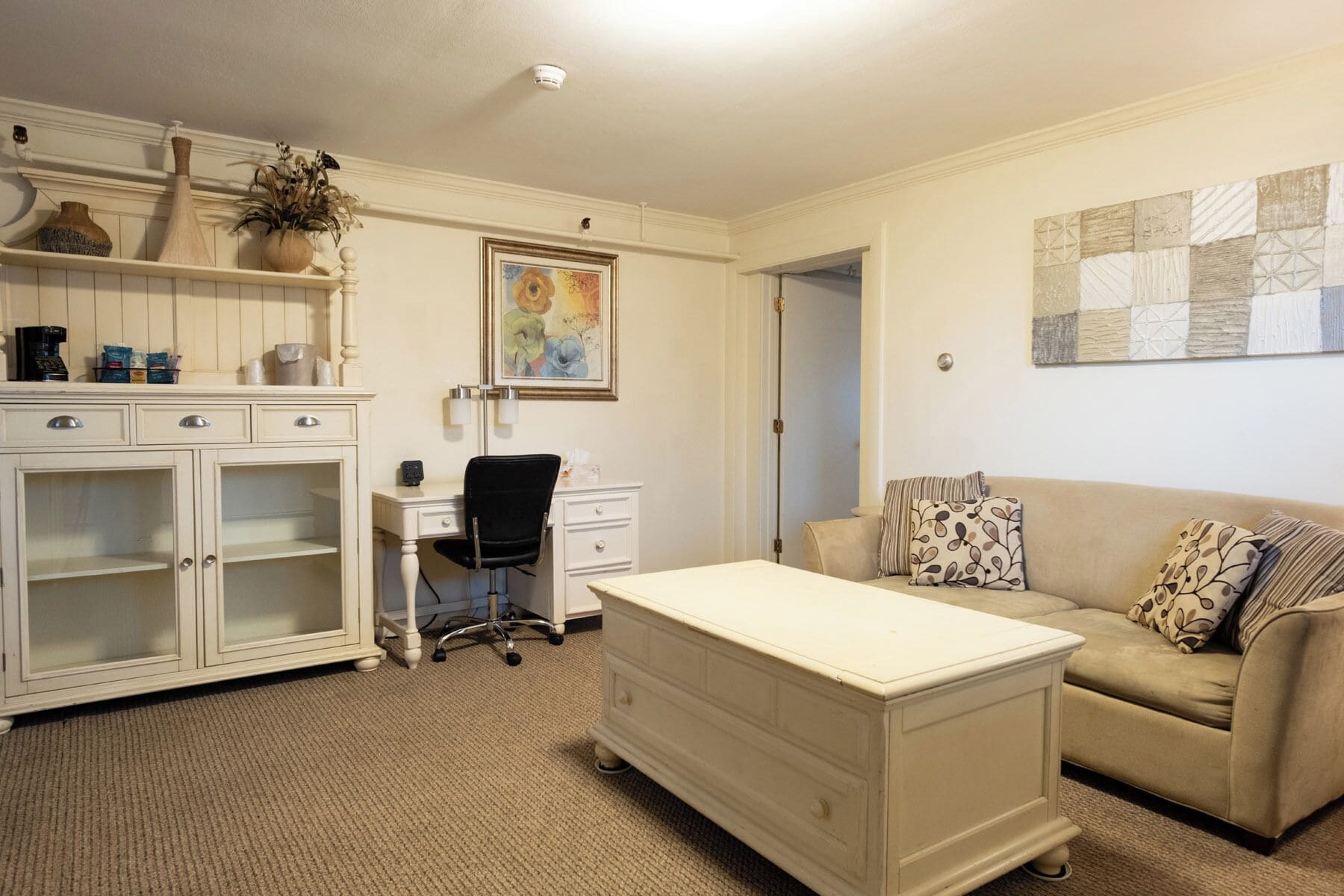 A well-lit room with a desk, a chair, a couch, and a cabinet. There is a painting on the wall and a plant on the shelf.