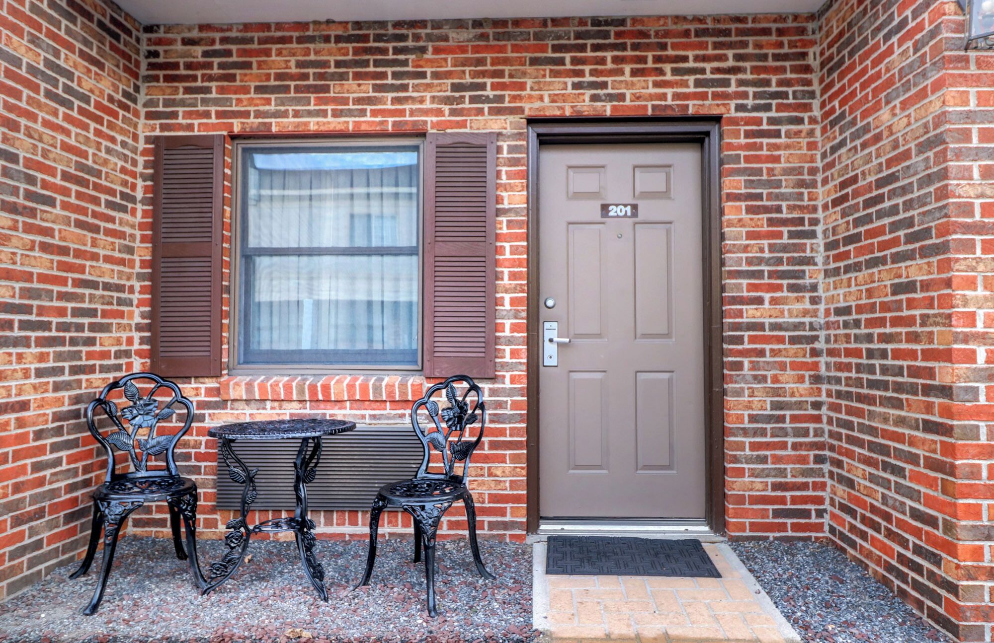 A brick building with a brown door marked '201', a window with brown shutters, and two ornate metal chairs with a small table in front.