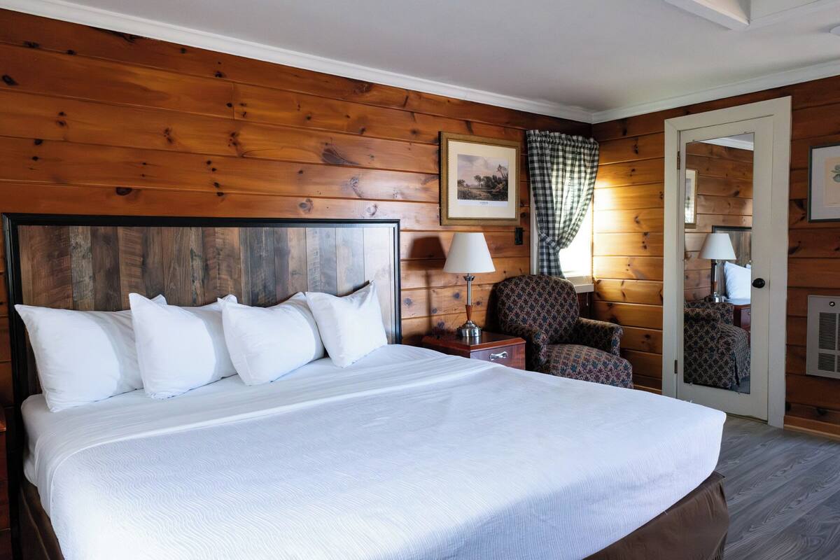 A wooden-paneled room with a large bed, a chair, a nightstand, and a door with a mirror.