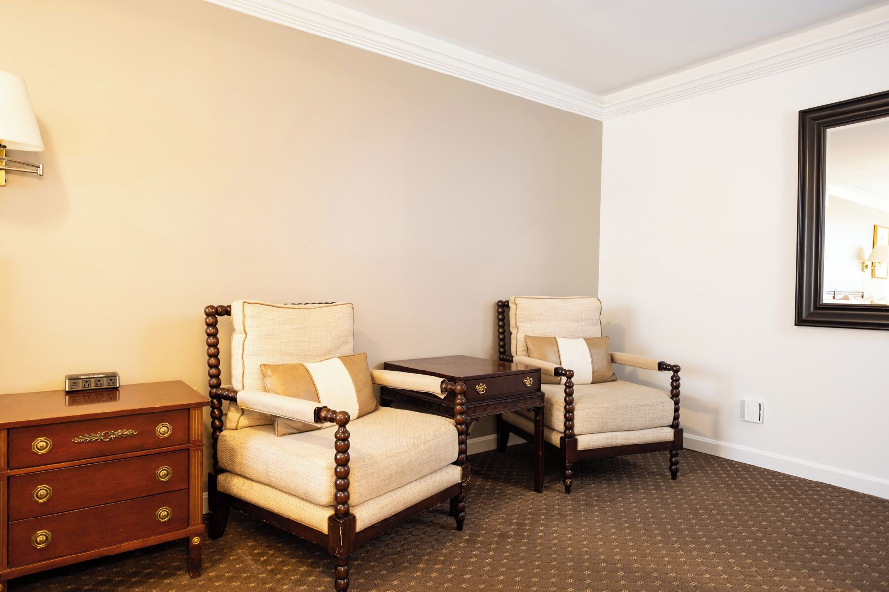 Une chambre d’hôtel avec deux fauteuils, une petite table en bois, une commode avec des tiroirs et une lampe fixée au mur.