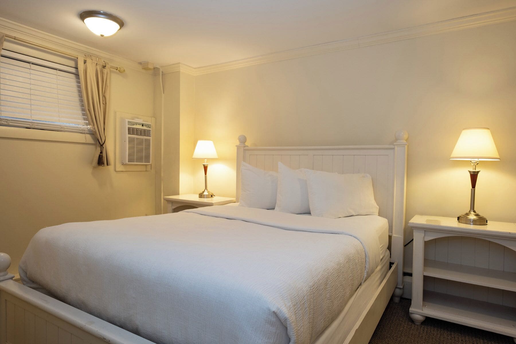 Un lit bien fait, avec du linge de lit blanc, deux tables de chevet avec des lampes, une fenêtre avec des stores et un climatiseur fixé au mur.